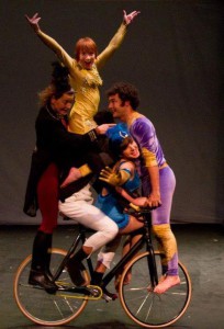 A tower of five circus performers - all balanced on a bike, with a woman in yellow on the top with her arms outstretched
