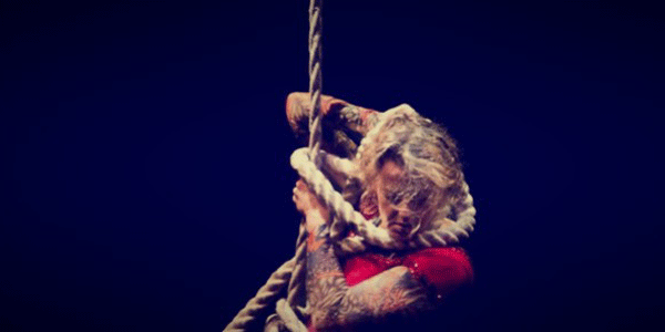 A photo of a woman's head and shoulders as she climbs a rope - she has loops of rope tangled around her tattooed arms and seems in a state of fury.