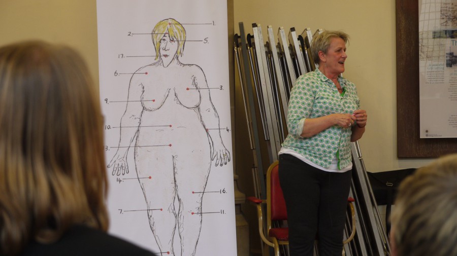 Bobby presenting about her show. She is next to a full sized drawing of herself, naked, with arrows pointing to parts of her body where she has been ill - her head, her breasts, her knees.