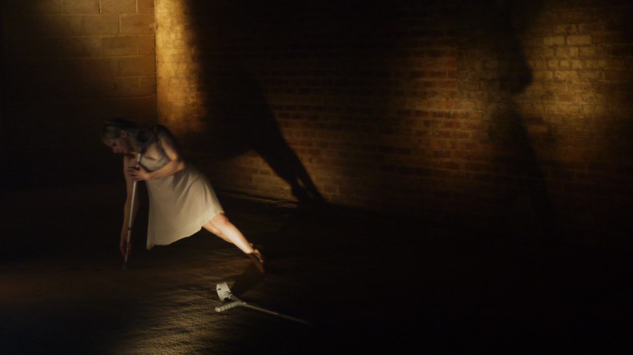 Claire Cunningham dancing with her crutches in a cellar with deep shadows. She is in a simple beige shift , one crutch is on the ground and she has her weight on the other with her body off to the right.