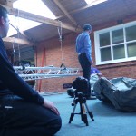 foreground - a camera man kneels on the floor, a camera on a tiny tripod inches high. Behind, Simon Mckeown unravels a grey deflated inflatable