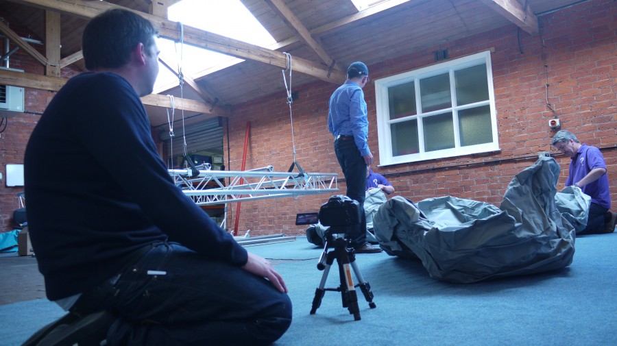 foreground - a camera man kneels on the floor, a camera on a tiny tripod inches high. Behind, Simon Mckeown unravels a grey deflated inflatable