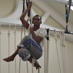 Ramesh Mayyappan up a rope - he is sitting holding the rope with his legs crossed in front of him.