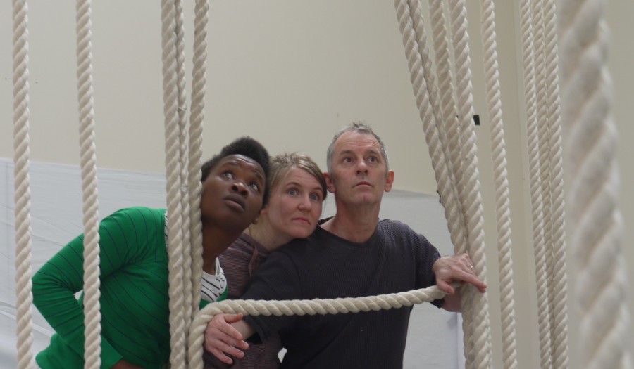The heads and shoulders of three actors can be seen - as though they are looking though a window (they have created a bottom frame for the window by holding up a piece of rope). 