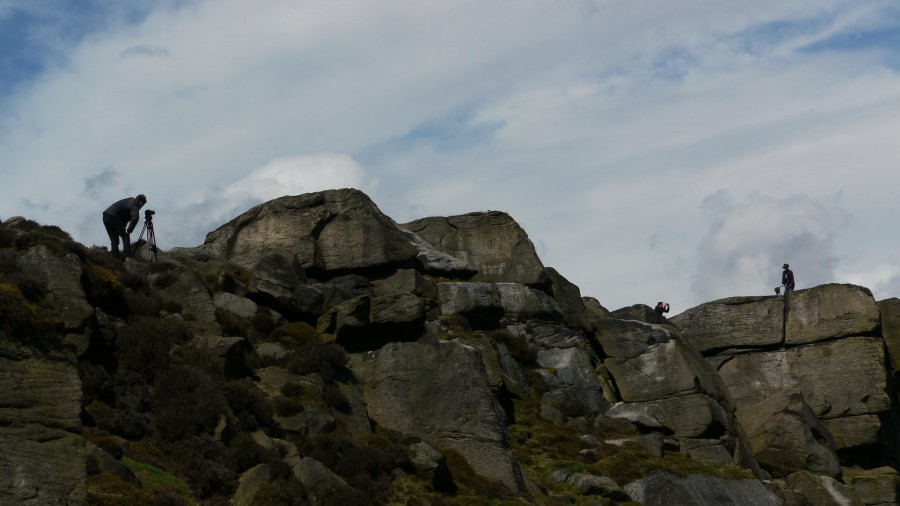 A huge rocky outcrop against a cloudy sky - two tiny figures can be seen - one filming, one in a stetson.