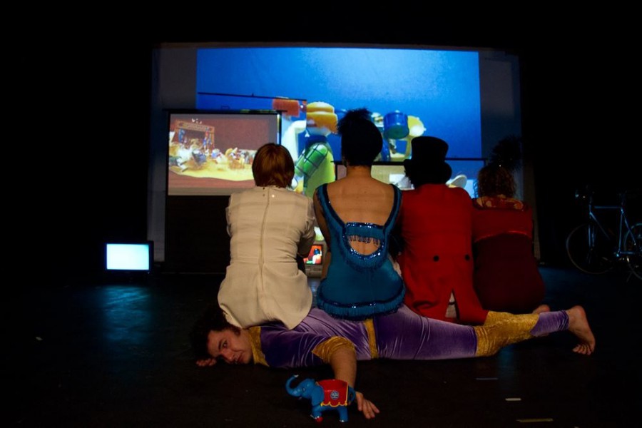 Three performers are watching a screen with their backs to us, they are dressed in circus clothes and are sitting on a fourth performer. The screen they are watching has a playmobil circus on it.