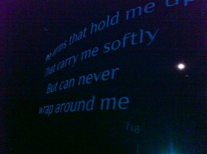 text on a gauze at the front of a stage - it reads the arms that hold me up, that carry me softly, but can never wrap around me. 