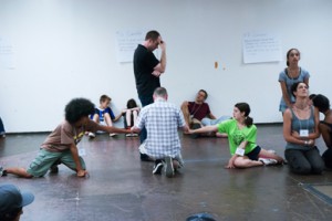image in a rehearsal room - one figure stands  his head in his hand and five others are on the floor around him forming a circle with their arms.