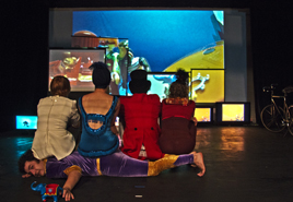the cast of Box of Frogs are sitting, backs to the audience, upon one of the performers - they are all watching a big screen upon which we can see circus Playmobile