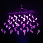 40+ people in white boiler suits are suspended in the air as though on a net. They are lit pink.