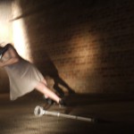 A still from the film of claire, dancing in a cellar with her crutches, one of which is on the floor in front of her out stretched leg