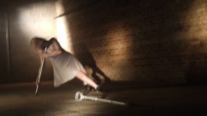 A still from the film of claire, dancing in a cellar with her crutches, one of which is on the floor in front of her out stretched leg