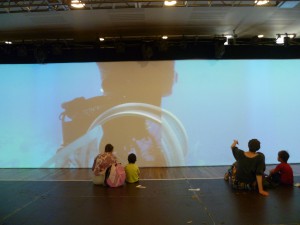 a wheelchair underwater can be seen projected on to a wall. two sets of families are sitting watching, pointing to what they see.