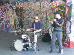 Jez playing a tin whistle next to a teenager playing a drum kit in front of a wall of graffiti